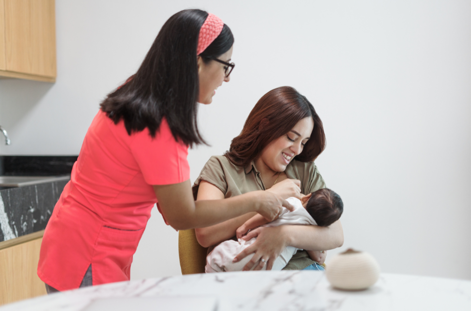 A woman wearing a pink headband and glasses looks on as another woman breastfeeds a baby.
